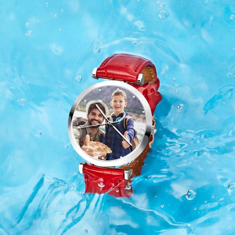 May include: A round wristwatch with a red leather band and a silver-colored case. The watch face displays a photograph of a man and a child. The watch is set against a blue water background.