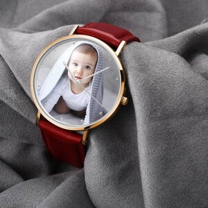 May include: A gold-toned wristwatch with a red leather band. The watch face features a photo of a baby. The watch has a round face with silver hands and hour markers. The watch is displayed on a gray fabric surface.