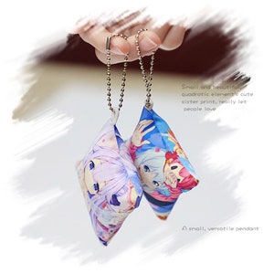 May include: Two small, square-shaped plush pillows with anime character designs. The pillows are attached to silver chains and are hanging from a hand.