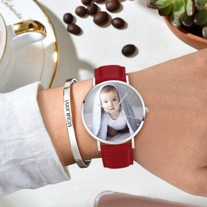 May include: A silver wristwatch with a red leather band features a custom photo of a baby. A silver bracelet with the inscription "MOMLXXVI" is also visible. The watch has silver hands and a silver case. The watch band is approximately 0.75 inches wide.