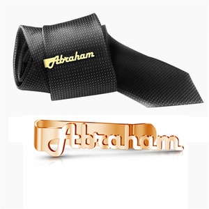 May include: Gold tie bar with the name "Abraham" engraved on it. The tie bar is shown on a black tie.