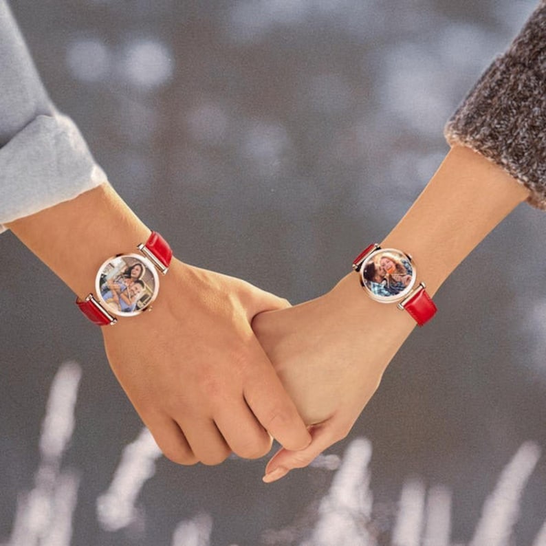 May include: Two wrists with matching watches. Each watch has a round face with a photo inside and a red leather band. The watches are held together, suggesting a connection or shared experience.