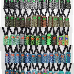 Puede incluir: Una colección de 60 cordones de paracord en varios colores y patrones. Los cordones se muestran en filas, mostrando una gama de diseños desde colores sólidos hasta tejidos intrincados. Cada cordón tiene un cordón negro para llevar al cuello.