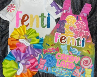 Op de afbeelding: Een regenboogkleurige strik met een wit T-shirt waarop "Fenti" in regenboogkleurige glitter staat en een spijkerbroek met een regenboogkleurige achtergrond en de woorden "Fenti Candy Land" in glitter.