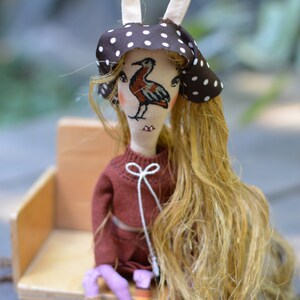 May include: A handmade doll with long blonde hair, wearing a brown and white polka dot hat and a maroon sweater. The doll's face has a bird design.