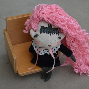 May include: A handmade doll with pink yarn hair, a black dress with white polka dots, and a bat design on its forehead. The doll is sitting on a wooden chair.