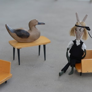 May include: A wooden duck figurine sits on a small wooden table with black legs. A doll with a black dress and a black headband sits on a small wooden chair with black legs.