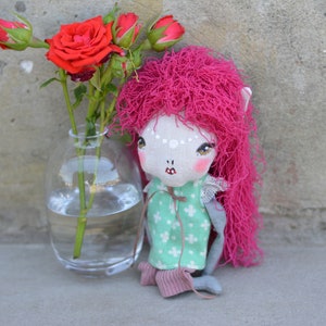 Könnte beinhalten: Eine handgemachte Puppe mit pinkfarbenem Garnhaar, einem grün-weiß gemusterten Kleid und grauen Hosen. Die Puppe sitzt auf einer grauen Oberfläche neben einer Glasvase mit roten Rosen.