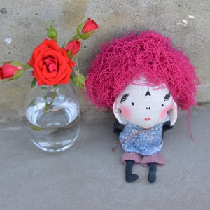 Könnte beinhalten: Eine handgemachte Puppe mit pinkfarbenem Garnhaar, einem weißen Gesicht und einem blau-weiß gemusterten Hemd. Die Puppe sitzt auf einer grauen Oberfläche neben einer durchsichtigen Glasvase mit roten Rosen.