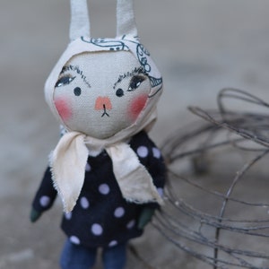 Könnte beinhalten: Eine handgefertigte Stoffpuppe mit einem weißen Gesicht, schwarzen Augen und rosa Wangen. Die Puppe trägt einen weißen Schal und ein schwarz-weißes gepunktetes Kleid. Die Puppe hat lange weiße Ohren und steht vor einem Drahtkäfig.