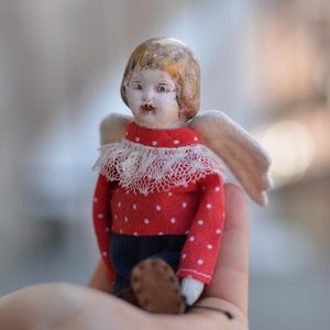 Könnte beinhalten: Eine kleine Porzellanpuppe mit einem rot-weiß gepunkteten Kleid und einem weißen Spitzenkragen. Die Puppe hat ein bemaltes Gesicht und braunes Haar. Die Puppe hält ein Paar Flügel.