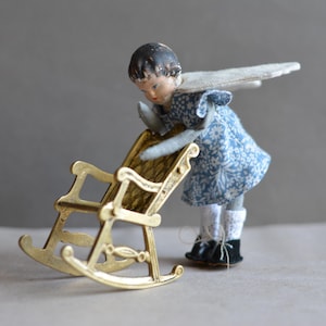 Könnte beinhalten: Eine kleine Puppe mit einem blau-weiß gemusterten Kleid und Flügeln sitzt in einem goldenen Schaukelstuhl. Die Puppe hat dunkles Haar und schaut nach unten.