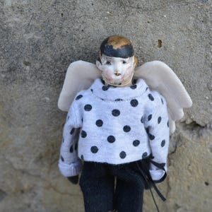 Könnte beinhalten: Eine Porzellanpuppe mit einem schwarz-weiß gepunkteten Pullover und einer schwarzen Hose. Die Puppe trägt ein Paar Engelsflügel.