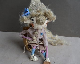 May include: A handmade doll with a long blonde wig, wearing a brown dress and blue socks. The doll is sitting in a wooden chair and has a blue flower in its hair.