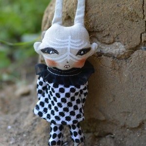 Könnte beinhalten: Eine handgemachte Puppe mit einem weißen Stoffkörper, einem schwarz-weiß gepunkteten Kleid und einem schwarzen und weißen Stoffkragen. Die Puppe hat ein bemaltes Gesicht mit schwarzen Augen, einer weißen Nase und einem roten Mund. Die Puppe steht neben einer rauen, braunen Wand.