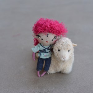 May include: A handmade doll with pink yarn hair and a blue and white floral print shirt is holding a white stuffed sheep.