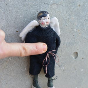 Könnte beinhalten: Eine kleine Porzellanpuppe mit bemalten Gesichtszügen, die einen schwarzen Pullover und eine Hose trägt und weiße Flügel hat. Die Puppe trägt braune Schuhe mit schwarzen Schnürsenkeln und steht auf einer grauen Oberfläche.