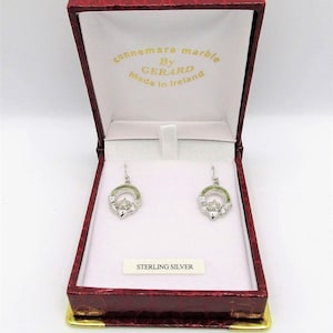 Può includere: Un paio di orecchini in argento sterling con un'intarsio in marmo verde Connemara a forma di anello Claddagh. Gli orecchini sono in una scatola rossa con le parole "Connemara Marble By Gerard Made in Ireland" all'interno del coperchio.
