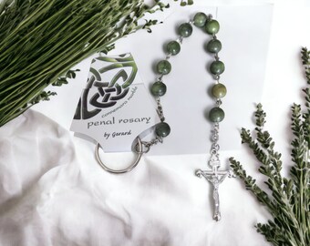 Puede incluir: Un rosario de mármol verde de Connemara con un dije de nudo celta de plata y un crucifijo de plata. El rosario está sobre una superficie blanca con follaje verde.