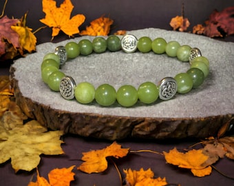 May include: A green jade bracelet with silver Celtic knot charms. The bracelet is on a wooden surface with autumn leaves.