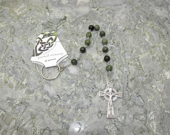 Puede incluir: Un rosario verde y negro con un colgante de cruz celta. El rosario está hecho de pequeñas cuentas redondas y tiene un anillo de plata unido a la parte superior. La tarjeta adjunta al rosario dice "penal rosary by Gerard".