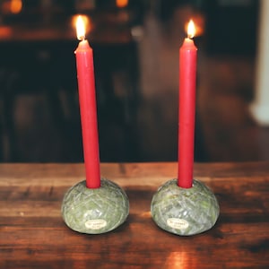 May include: Two red candles are lit and placed in green marble candle holders. The candle holders have a circular base and a small opening in the center to hold the candle.