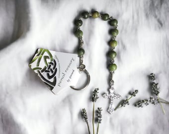 Puede incluir: Un rosario de piedra verde con un dije de nudo celta de plata y una cruz de plata. El rosario está sobre un fondo de tela blanca.