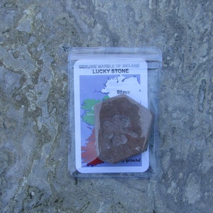 May include: A brown, pentagonal stone with an embossed four-leaf clover design, enclosed in a clear plastic pouch. The packaging features a card labeled "GENUINE MARBLE OF IRELAND" and "LUCKY STONE" with a map of Ireland highlighting the province of Ulster.