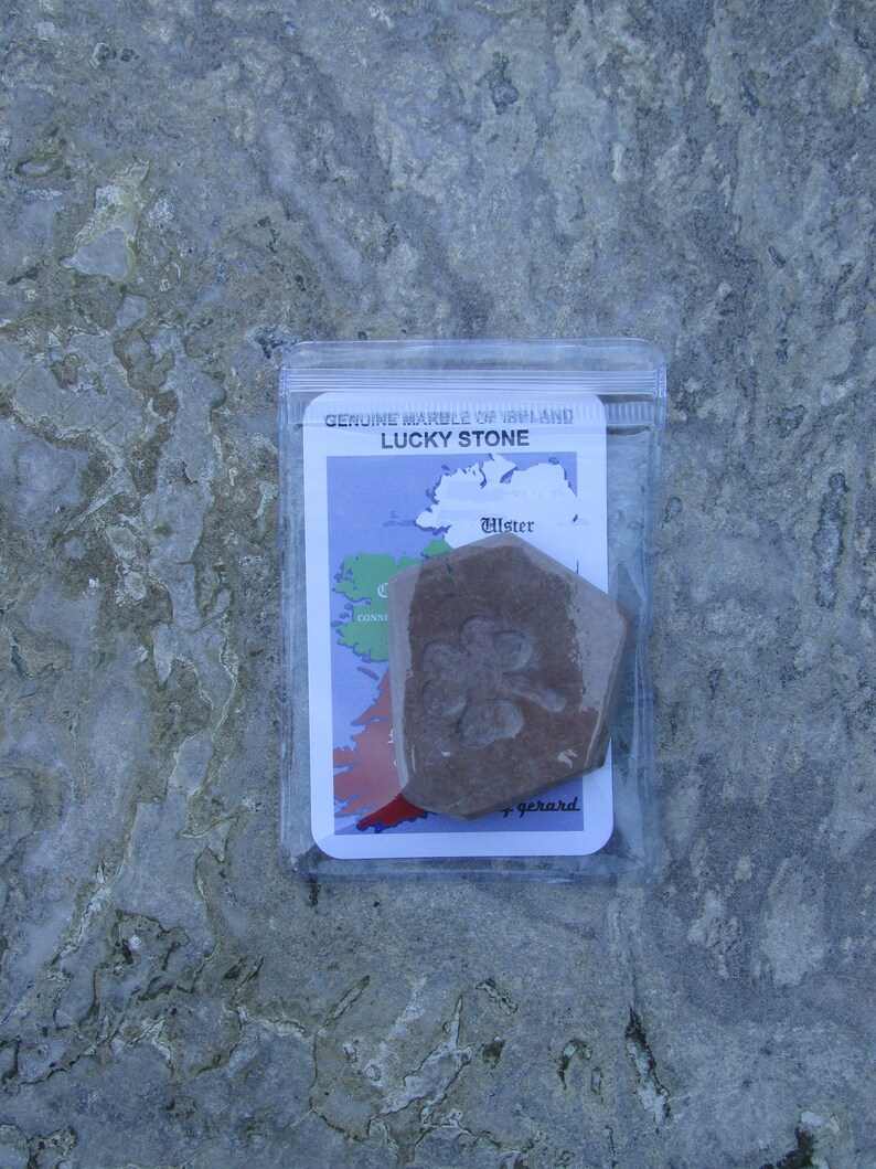 May include: A brown, pentagonal stone with an embossed four-leaf clover design, enclosed in a clear plastic pouch. The packaging features a card labeled "GENUINE MARBLE OF IRELAND" and "LUCKY STONE" with a map of Ireland highlighting the province of Ulster.