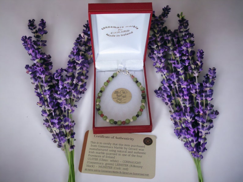 May include: A red box with a green and red Connemara marble bracelet inside. The box is open and the bracelet is visible. The box is surrounded by lavender flowers.