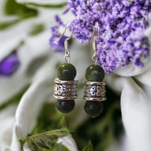 May include: A pair of dangle earrings featuring dark green, round beads. The earrings have silver-colored metal accents with a Celtic knot design. The earrings are displayed against a backdrop of white and purple flowers.
