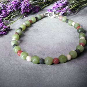 May include: A green, red, white, and black beaded necklace with a silver clasp. The necklace is made of natural stones and is laid out on a gray surface.