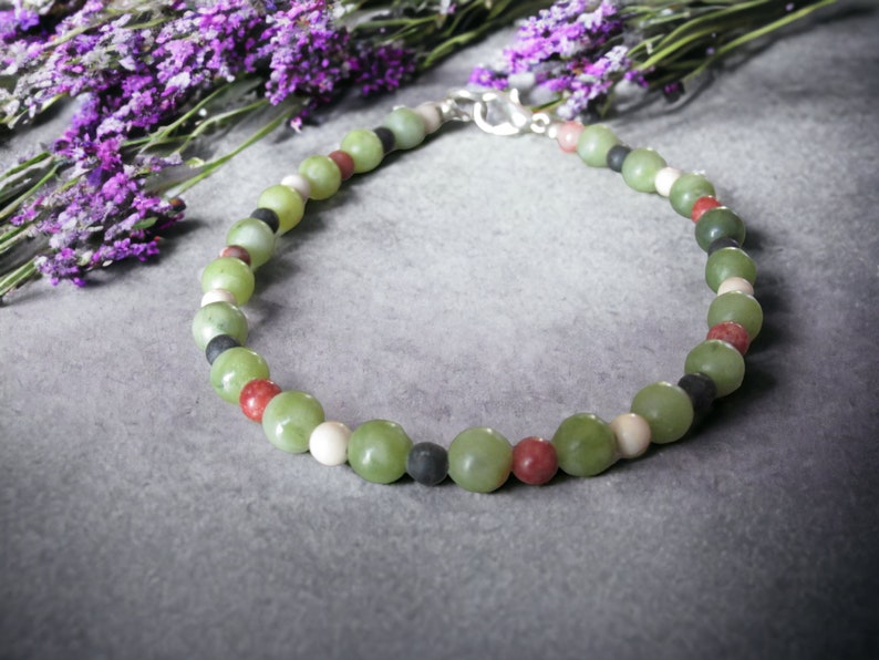 May include: A green, red, white, and black beaded necklace with a silver clasp. The necklace is made of natural stones and is laid out on a gray surface.