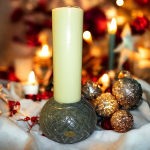 May include: A green stone candle holder with a carved design holds a tall, white pillar candle. The candle holder is surrounded by small, gold-colored ornaments.