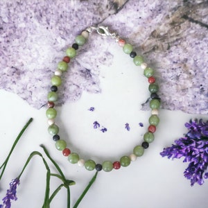 May include: A green, red, white, and black beaded bracelet with a silver clasp. The bracelet is on a white surface with lavender flowers.