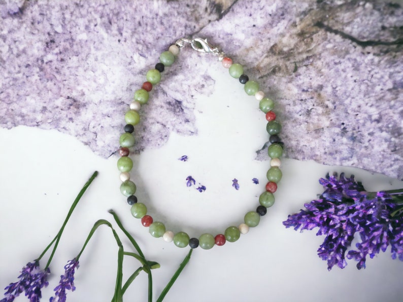 May include: A green, red, white, and black beaded bracelet with a silver clasp. The bracelet is on a white surface with lavender flowers.