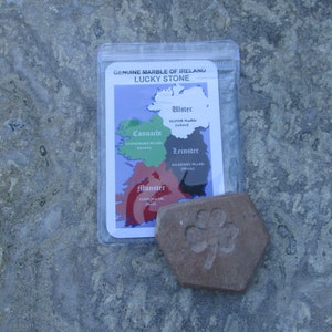 May include: A reddish-brown hexagonal stone featuring an embossed shamrock design, accompanied by a clear plastic pouch containing an informational card titled GENUINE MARBLE OF IRELAND LUCKY STONE, which displays a map of Ireland with four regions.