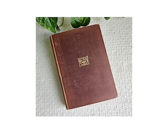 May include: A brown hardback book with a gold design on the cover. The book is closed and lying flat on a white surface.