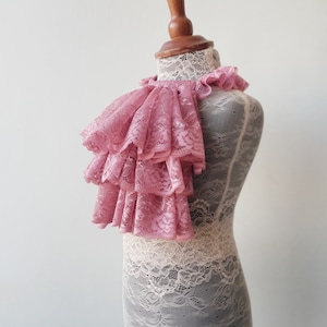 May include: A pink lace ruffle collar with multiple layers. The collar is displayed on a white lace-covered mannequin.