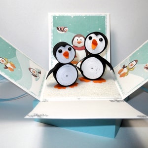 Może przedstawiać: Kartka z wyskakującym obrazkiem przedstawiająca zimową scenę z trzema pingwinami wykonanymi z papierowych rolek. Kartka jest otwarta, a pingwiny stoją na białej, błyszczącej powierzchni. Kartka jest ozdobiona niebieską wstążką.
