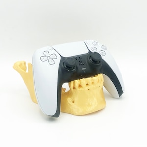 May include: A white and black PlayStation 5 controller rests on a tan-colored jawbone model. The controller has a black center section with buttons and a PlayStation logo. The jawbone model is detailed with teeth.