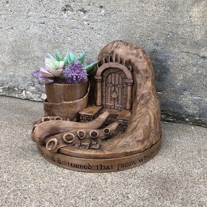 May include: A wooden planter shaped like a cave with a door and an octopus tentacle base. The planter has a succulent plant and a purple flower inside. The base of the planter has the text "and so disturbed that from which t"