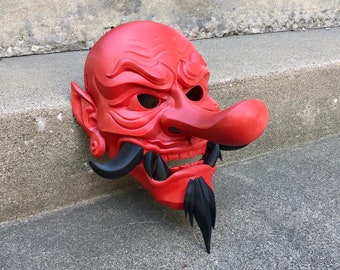 Könnte beinhalten: Eine rote und schwarze japanische Oni-Maske mit einer großen, übertriebenen Nase. Die Maske ist mit komplizierten Details geschnitzt und hat einen grimmigen Ausdruck.