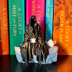 Puede incluir: Una figura impresa en 3D de color negro de una figura encapuchada sentada en un banco de piedra, sosteniendo una taza de té y un platillo. La figura está sentada frente a una estantería con varios libros titulados "A Court of..." visibles.