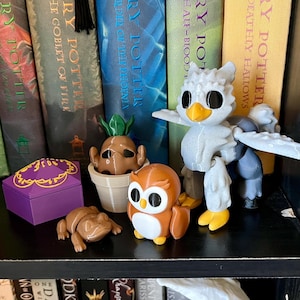 May include: A collection of Harry Potter themed figurines on a bookshelf. The figurines include a brown mandrake plant, a purple box, a brown frog, an orange owl, and a white and gray bird.