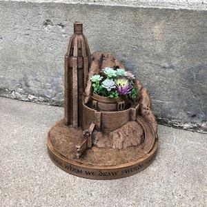 May include: A brown, 3D printed planter shaped like a castle with a tower and a quote that reads "Fear when we draw swords from the stones". The planter is filled with green and purple succulents.