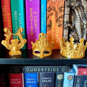 May include: Gold-colored decorative items, including a lyre, a masquerade mask, and a crown, are displayed on a black shelf in front of a row of books. The books have colorful spines and titles like "A Court of Thorns and Roses."