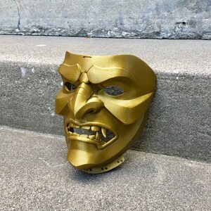 May include: A gold-coloured, detailed, plastic mask with a fierce expression. The mask has a wide mouth with sharp teeth, a prominent nose, and deep-set eyes.