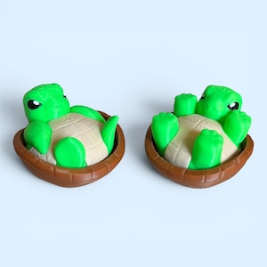 May include: Two green turtle figurines with tan shells and brown baskets. The turtles are lying on their backs with their limbs extended. The figurines have large, black eyes and are set against a light blue background.