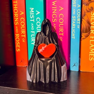 Op de afbeelding: Een zwart beeldje van een figuur met een kap die een feloranje hart met een roze strik vasthoudt. Het beeldje staat voor een rij kleurrijke boeken met titels als "A Court of Thorns and Roses" en "A Court of Mist and Fury".